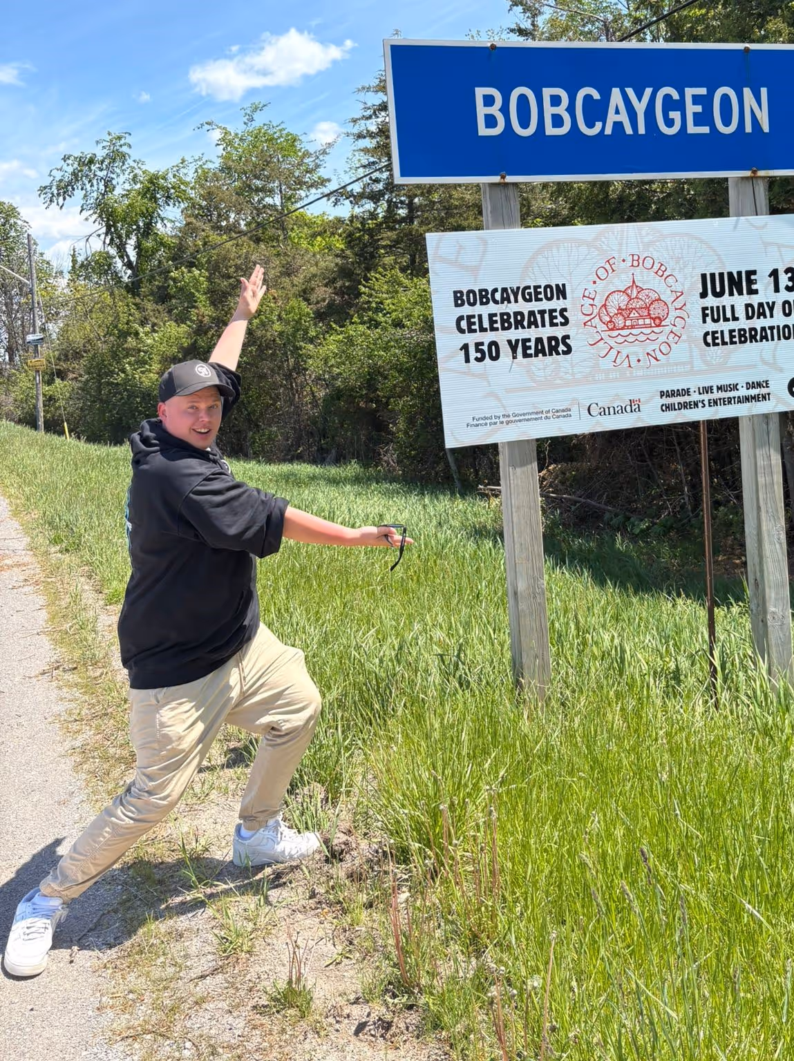 Person posing excitedly beside a Bobcaygeon sign and a banner about its 150-year celebration.