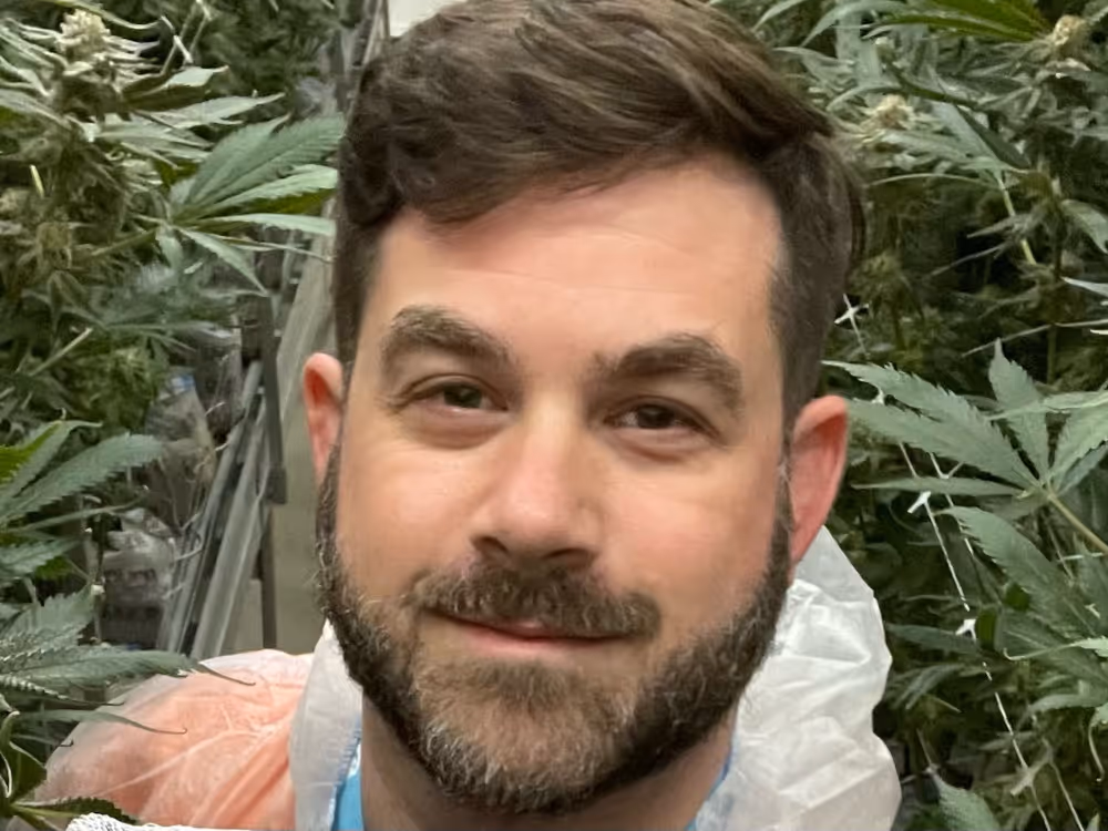 Smiling man with beard among tall green plants in an indoor garden or greenhouse.