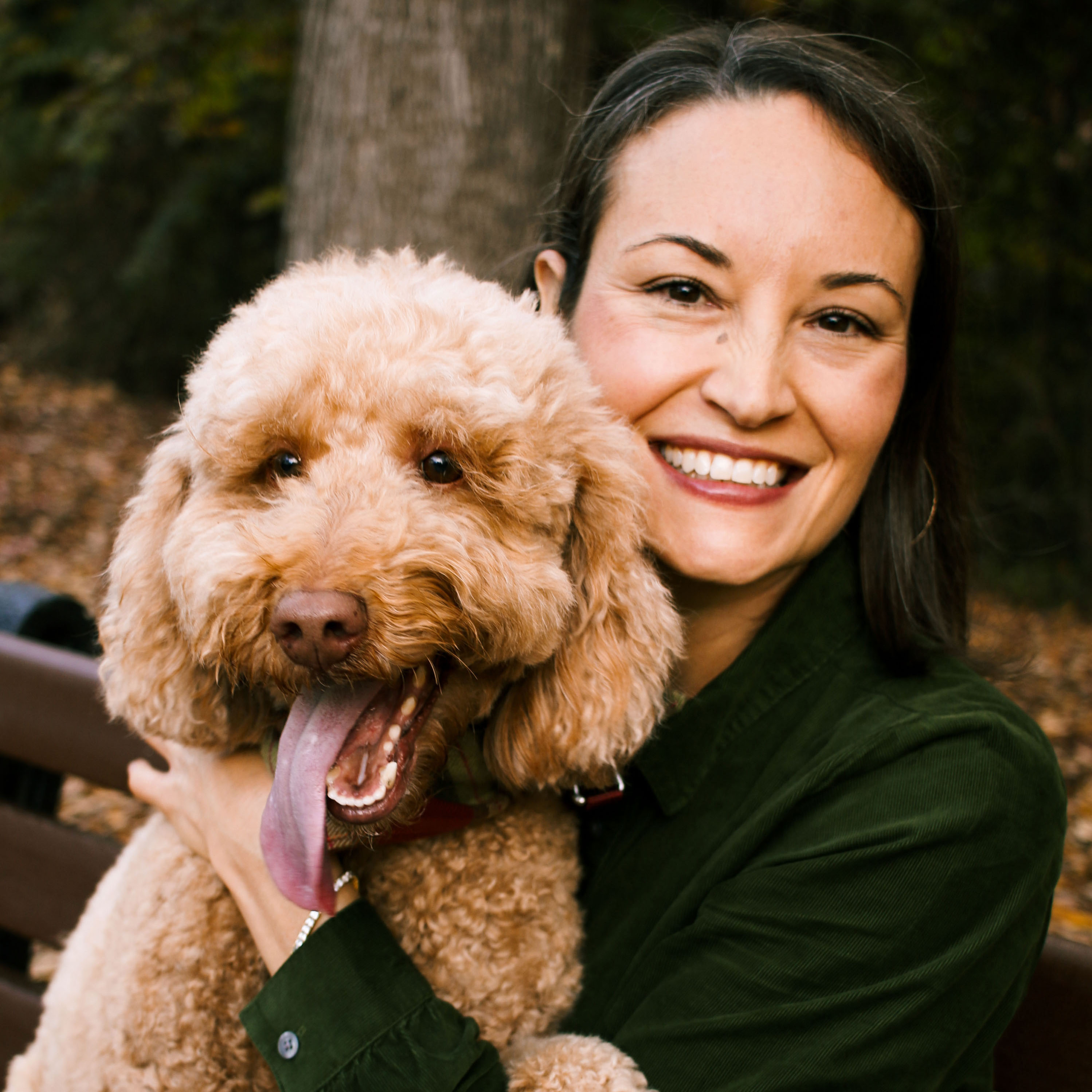 A young woman with brown hair holds a light brown medium-sized poodle in her lap while sitting on a park bench.