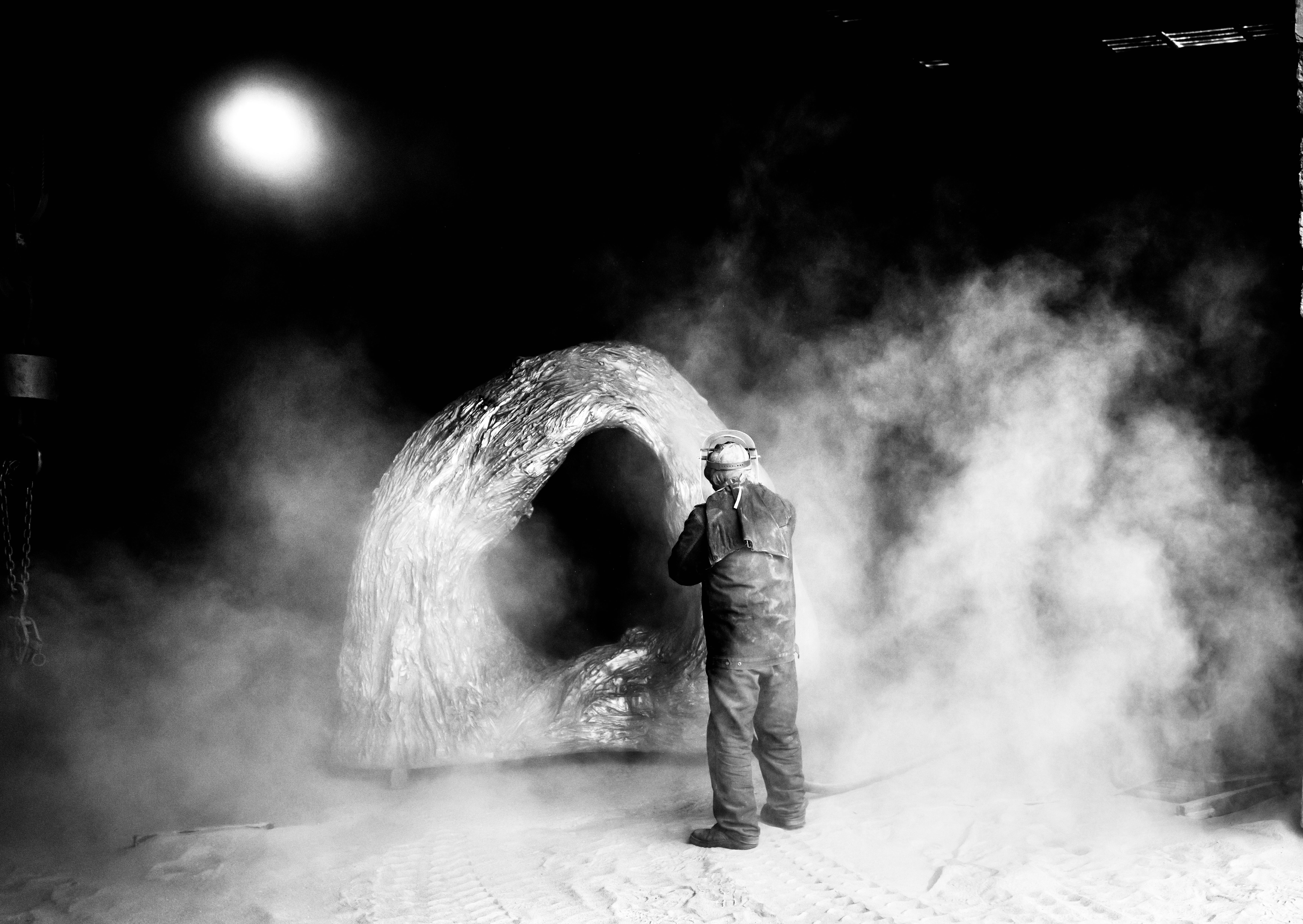 Worker in protective gear standing amid smoke while shaping a large metal sculpture.