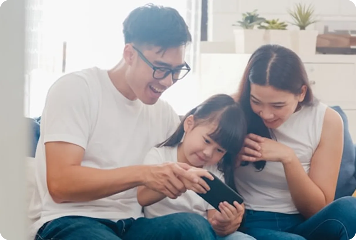Smiling Asian family of father, mother, and daughter sitting on a couch looking at a smartphone together.