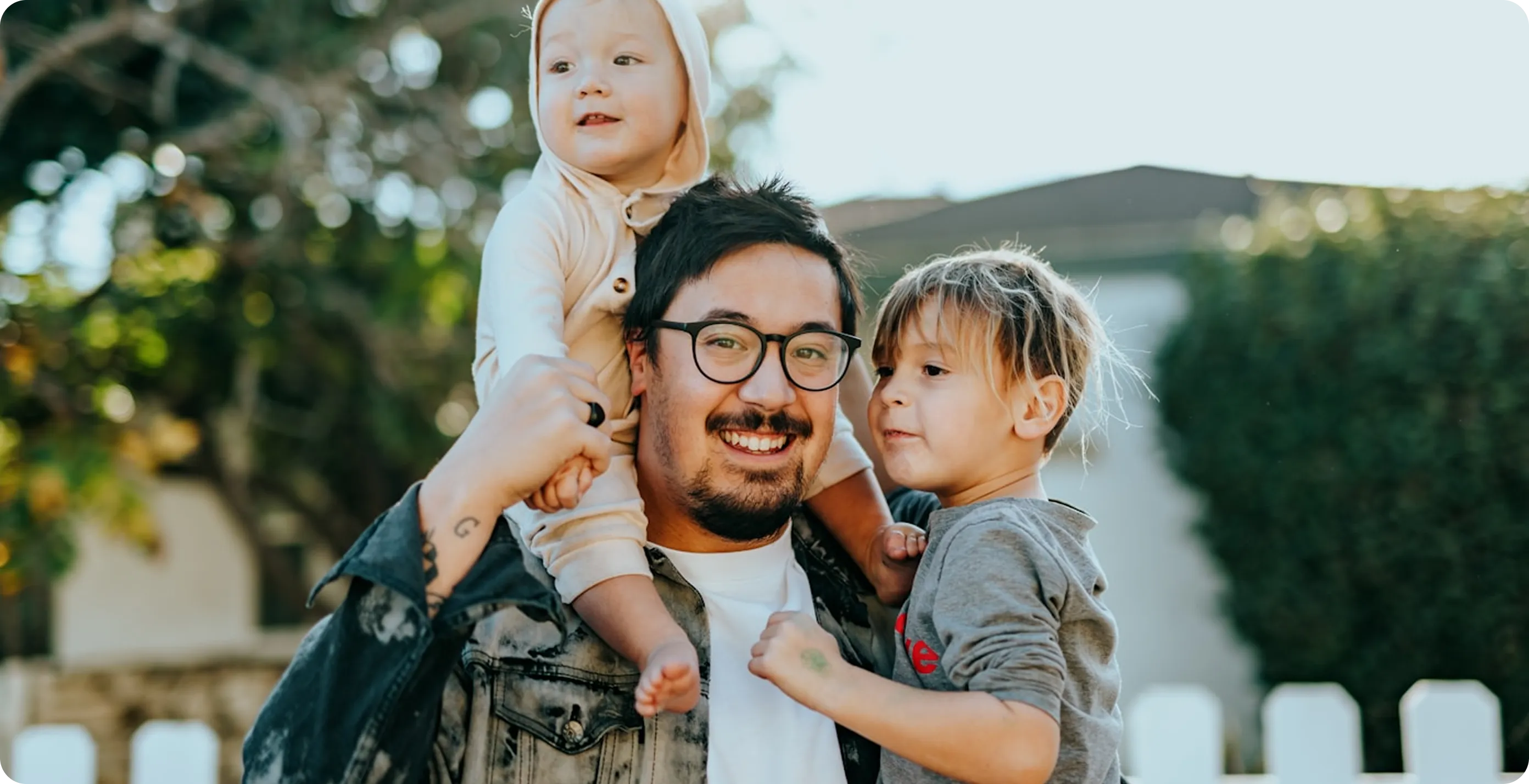 Smiling man wearing glasses holding two young children outdoors near a white fence and greenery.