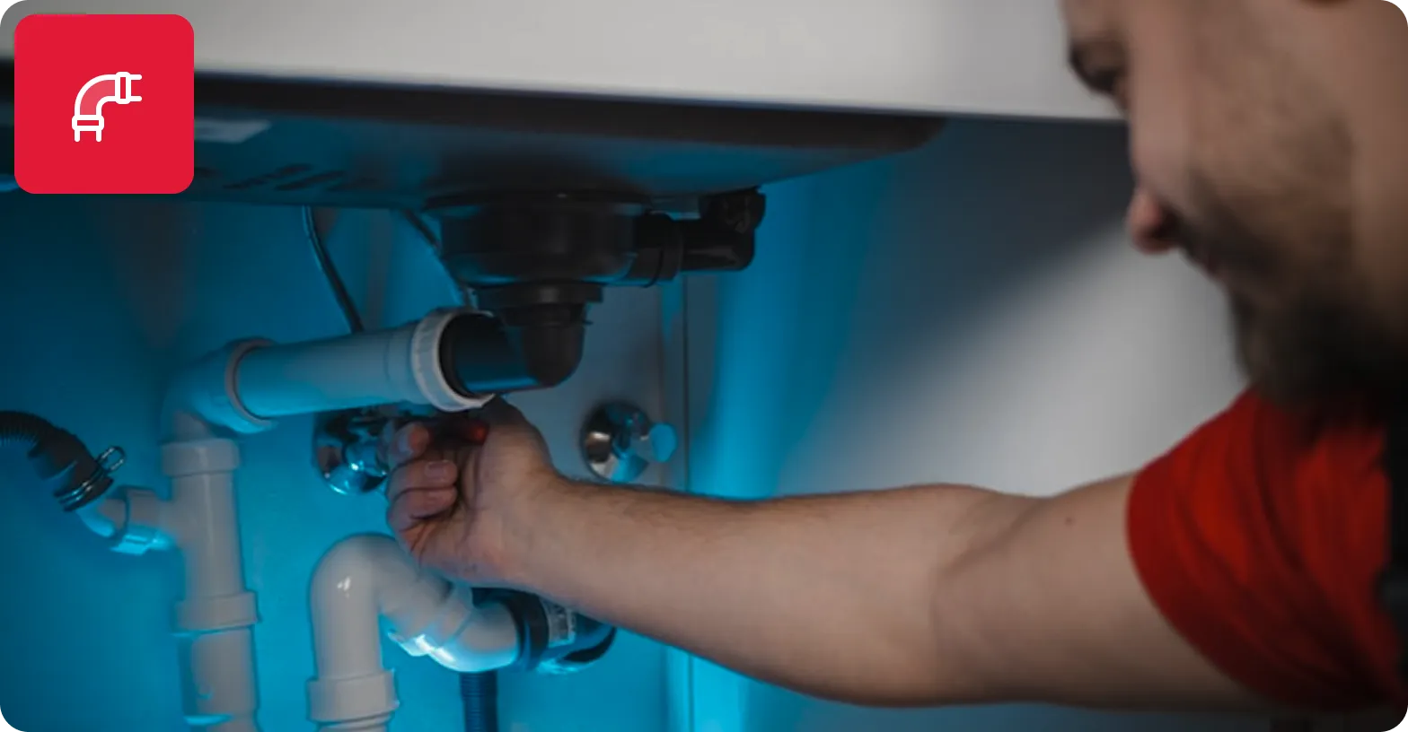 Man in red shirt fixing plumbing pipes under a sink.