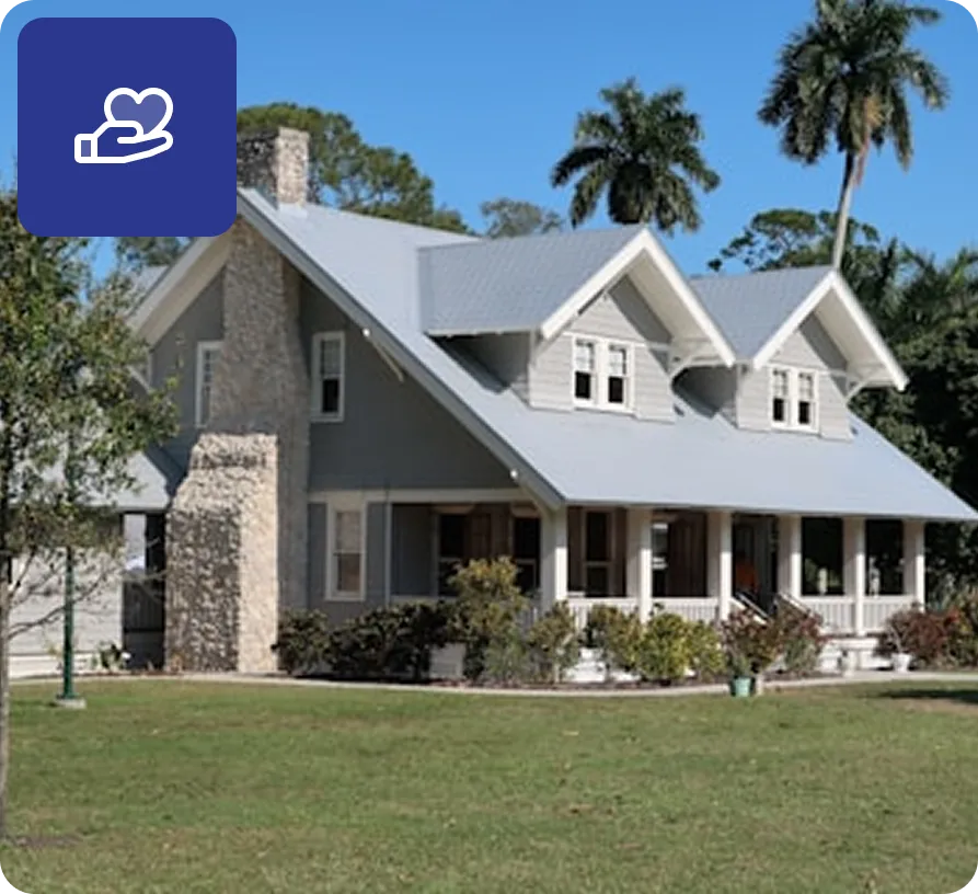 Large gray house with a stone chimney, white porch columns, and two dormer windows, surrounded by palm trees and a green lawn.