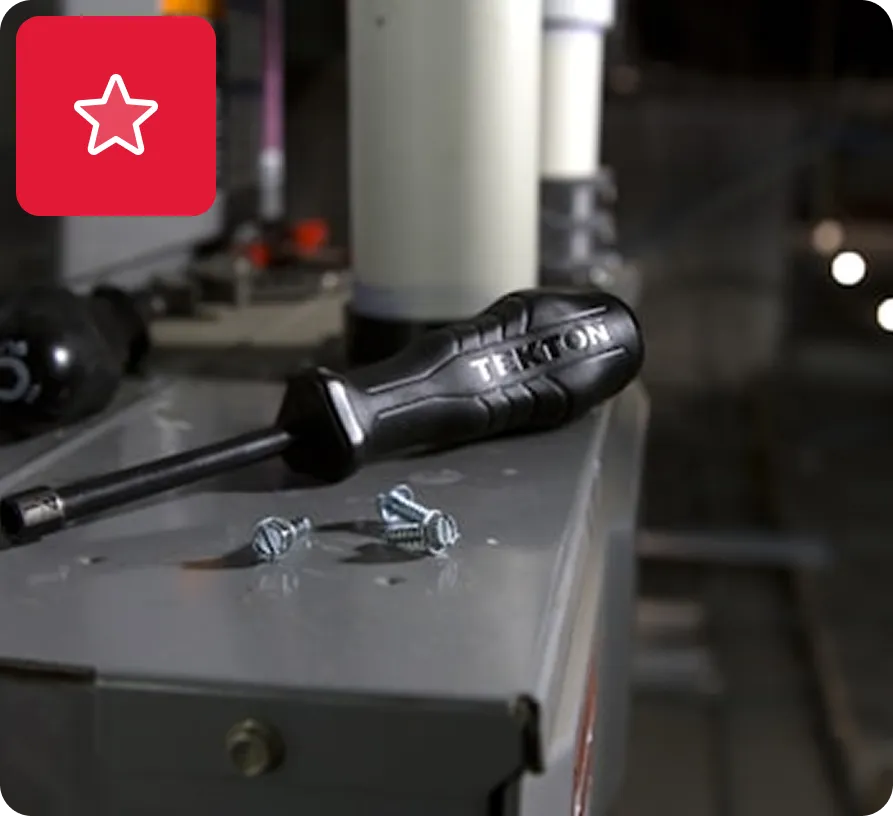 Black Tekton screwdriver lying on a metal surface next to three screws in a workshop setting.