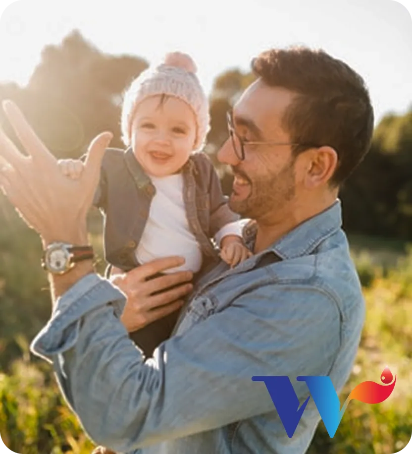 Smiling man wearing glasses and denim jacket holding a happy baby in a knit hat outdoors during golden hour.