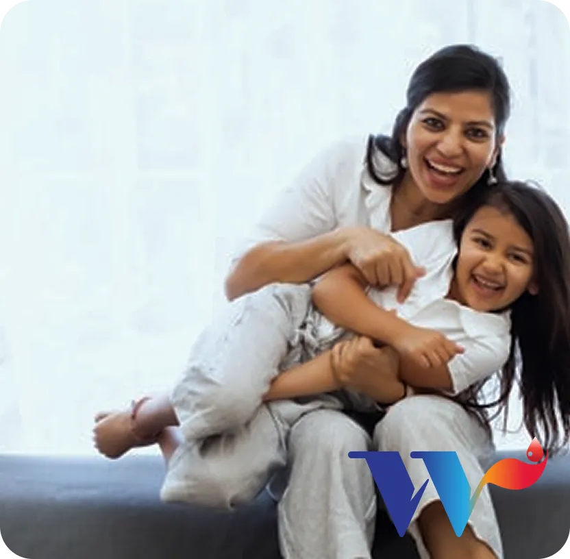 Smiling woman playfully holding and hugging a happy young girl on a couch in a bright room.