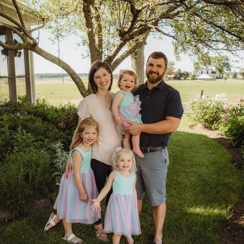 Smiling family of five in a garden with green grass and trees, including a man holding a baby and three young girls in matching pastel dresses.