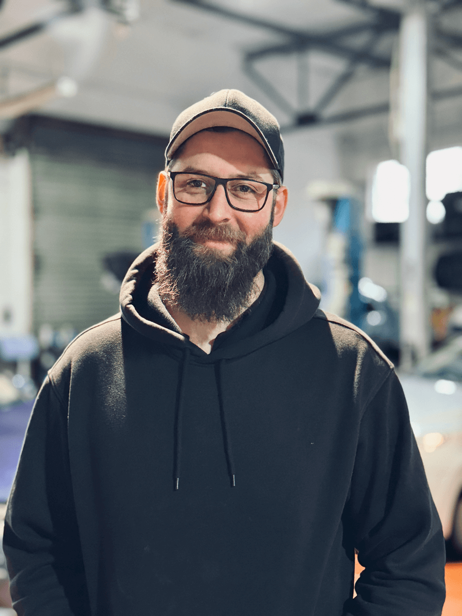 Bärtiger Mann mit Brille, schwarzem Kapuzenpulli und schwarzer Mütze, der in einer Werkstatt oder Garage lächelt.