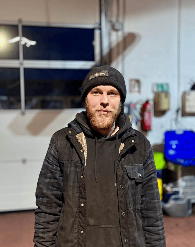 Bärtiger Mann mit schwarzer Puma-Mütze und dunkler Kapuzenjacke, der in einem Innenraum vor einem industriellen Hintergrund steht.