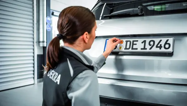 Person in DEKRA-Weste bringt in einer Garage einen Aufkleber auf dem Nummernschild eines Autos an.