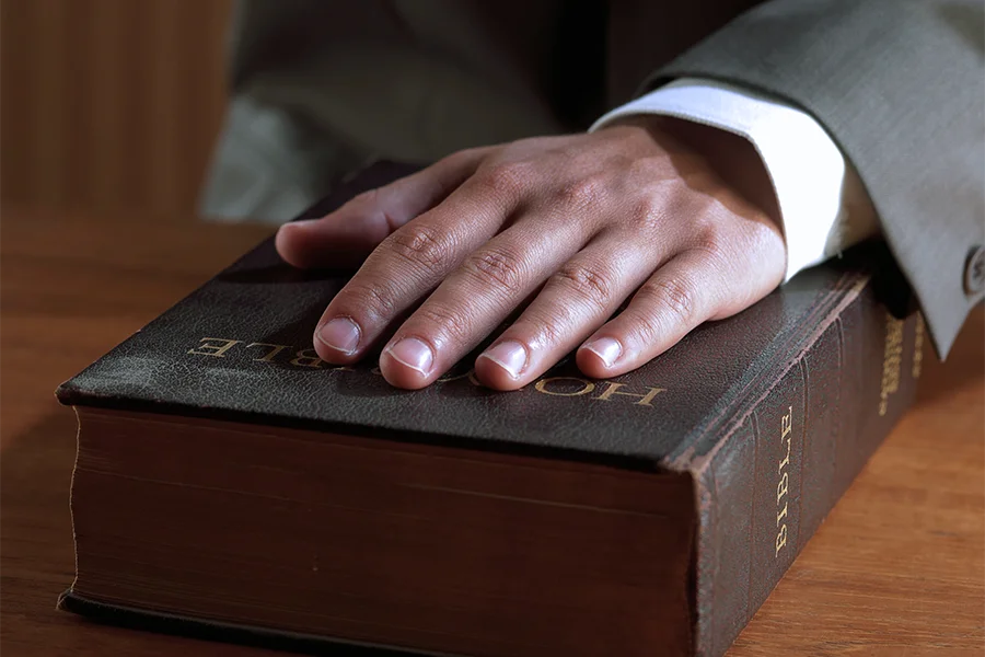 A hand in a suit resting on a closed Bible on a wooden surface.