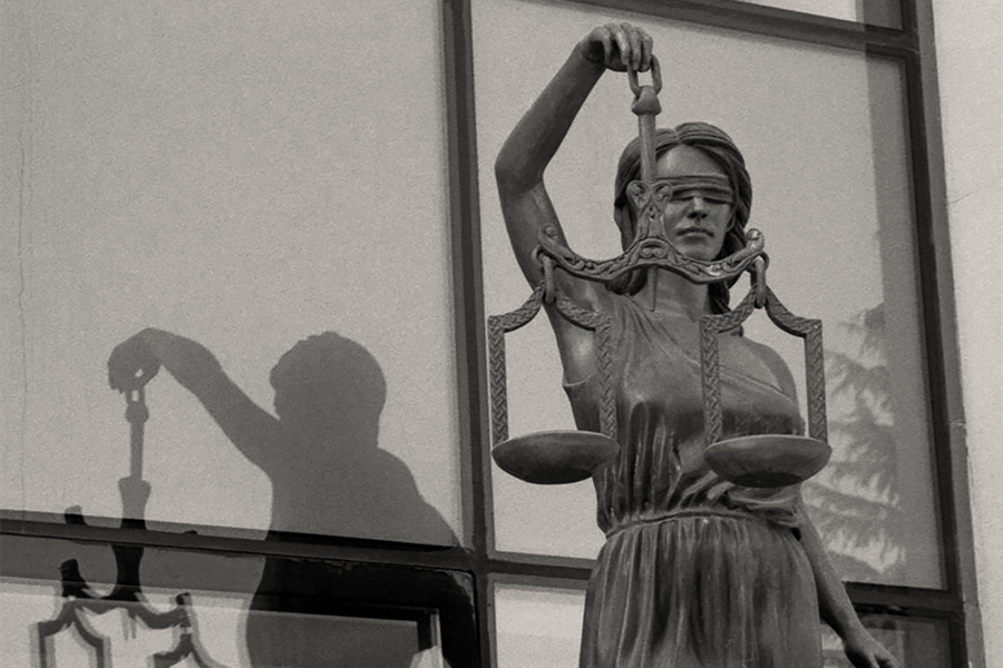 Blindfolded Lady Justice statue holding scales in front of glass window with her shadow reflected on it.