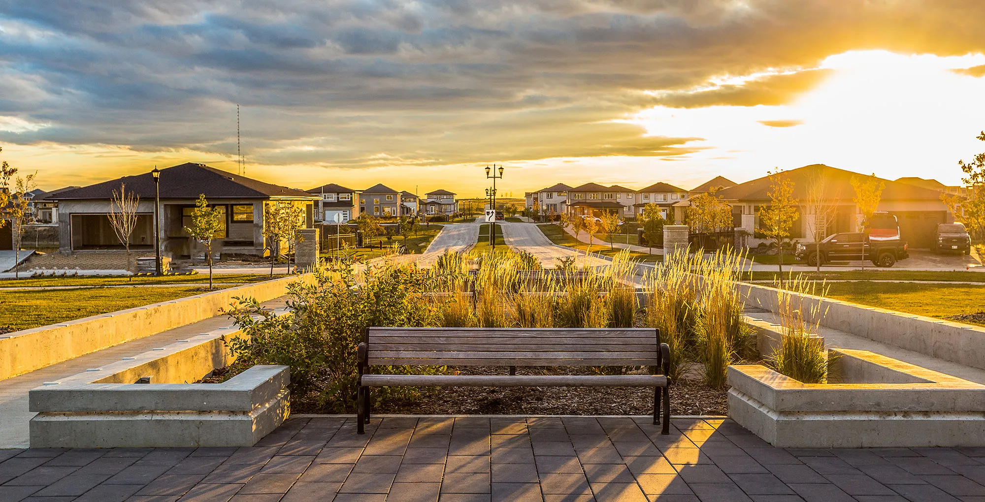 A scenic sunset view of a suburban neighborhood with a paved road leading to rows of modern houses, a wooden bench in the foreground, and soft golden light illuminating the landscape.