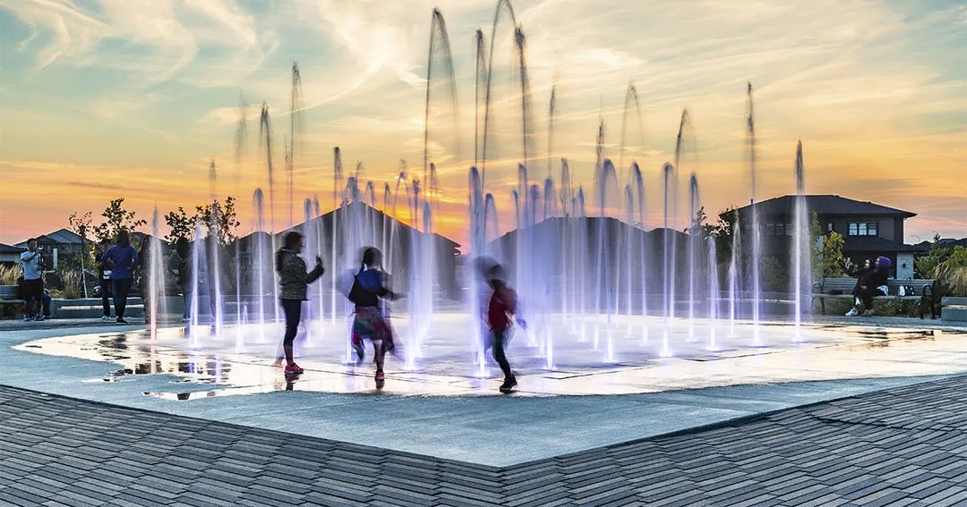 Children playing in an interactive water fountain at sunset, with illuminated jets of water creating a dynamic and colorful scene.