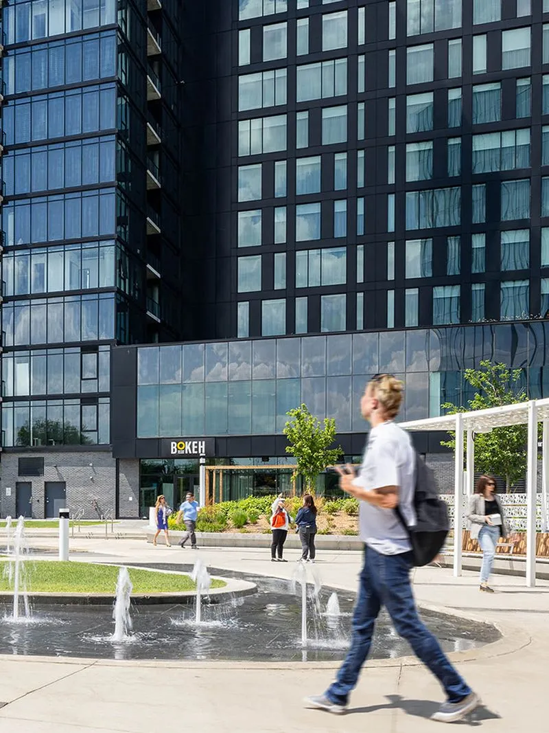 A pedestrian-friendly urban plaza featuring a water fountain, greenery, and people walking, set against the backdrop of a contemporary high-rise building with reflective glass windows.