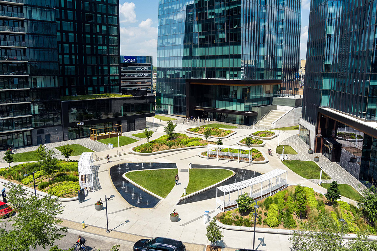 Modern outdoor seating area with two cushioned sofas facing a wooden coffee table under a pergola, overlooking a pool surrounded by tropical plants and lounge chairs.