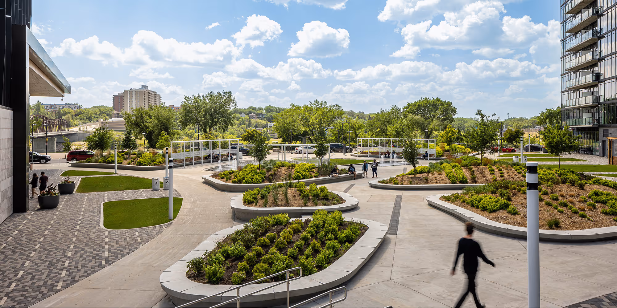 Modern mixed-use development with glass office towers surrounding a central plaza featuring landscaped gardens, water features, and pedestrian walkways.