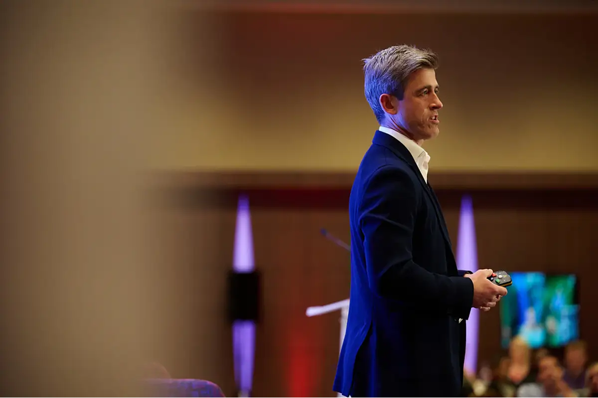 Scott Walker speaking and holding a remote control at an event with blurred audience in the background.