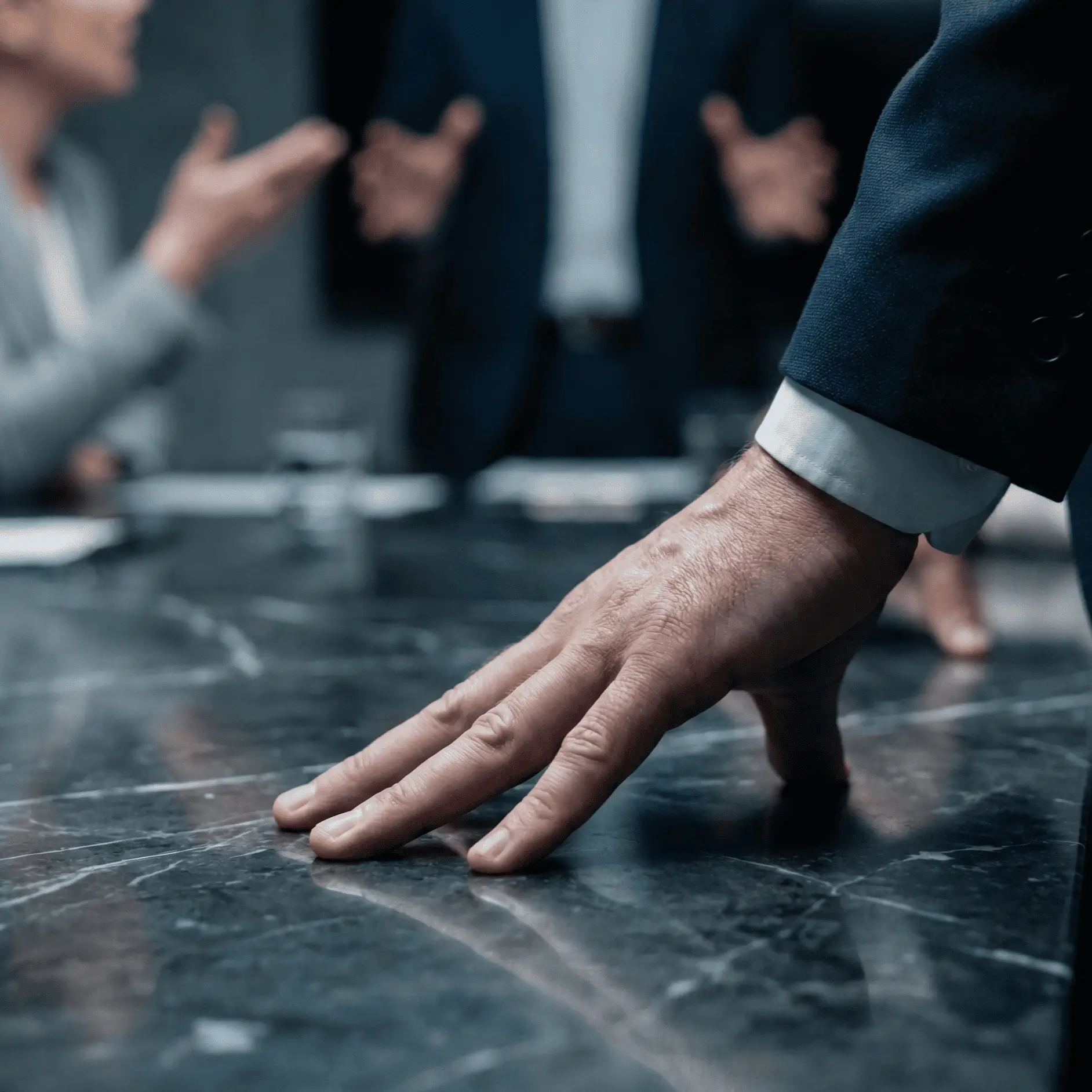 Man's hand pressing firmly on a marble table during a tense business meeting with blurred people gesturing in the background.