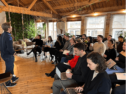 Scott Walker as speaker presenting to an attentive audience seated in a wooden loft-style room with plants and large windows.