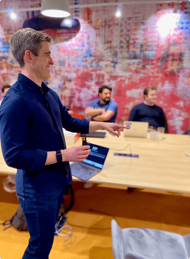 Scott Walker holding a clicker and pointing while speaking to two seated men in a conference room with a large wooden table and colorful abstract wall art.