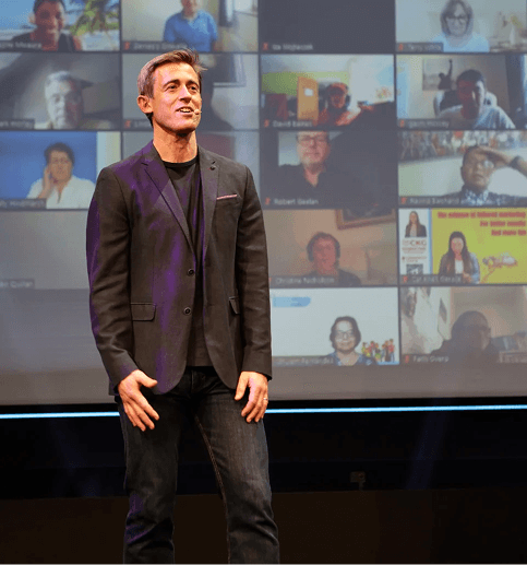 Scott Walker speaking on stage with a large screen showing multiple video call participants behind him.
