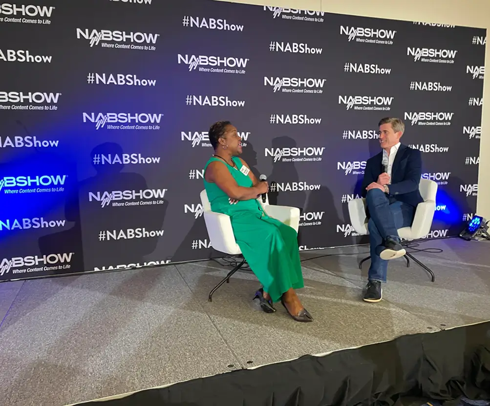 Two people seated on stage, one of it is Scott Walker, they having a discussion at the NAB Show event, with a black backdrop featuring repeated #NABShow logos.