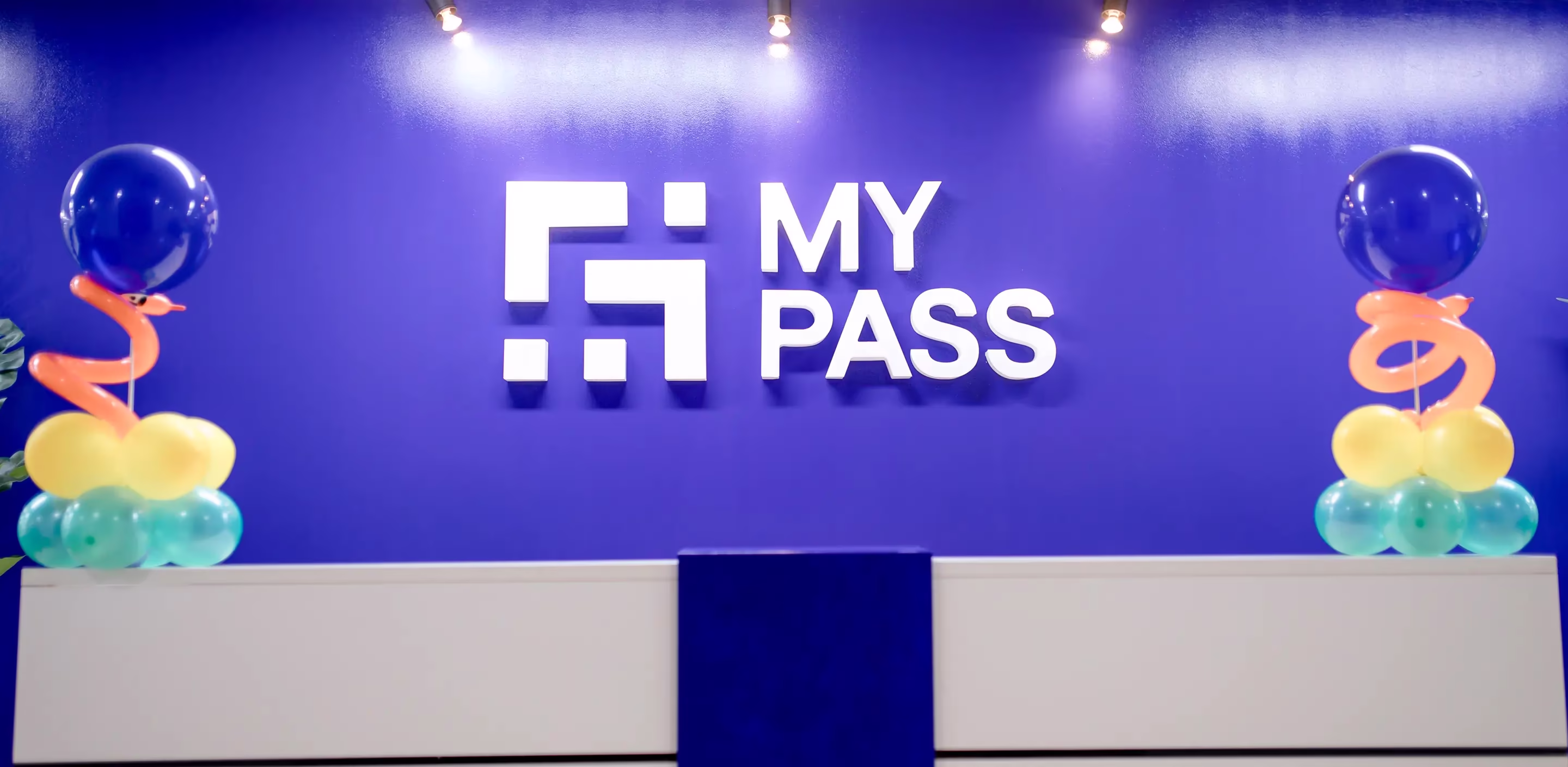Mostrador de recepción con una pared azul brillante detrás en la que se muestra el logotipo blanco de MY PASS y decoraciones con globos de colores a ambos lados.