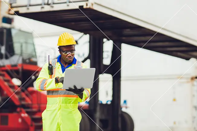 Trabajador de la construcción con equipo de seguridad de alta visibilidad y casco utilizando un ordenador portátil en un sitio de trabajo industrial.