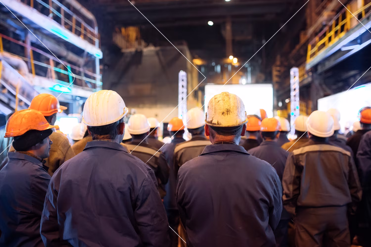 Grupo de trabajadores industriales con cascos blancos y naranjas viendo una presentación en una fábrica.