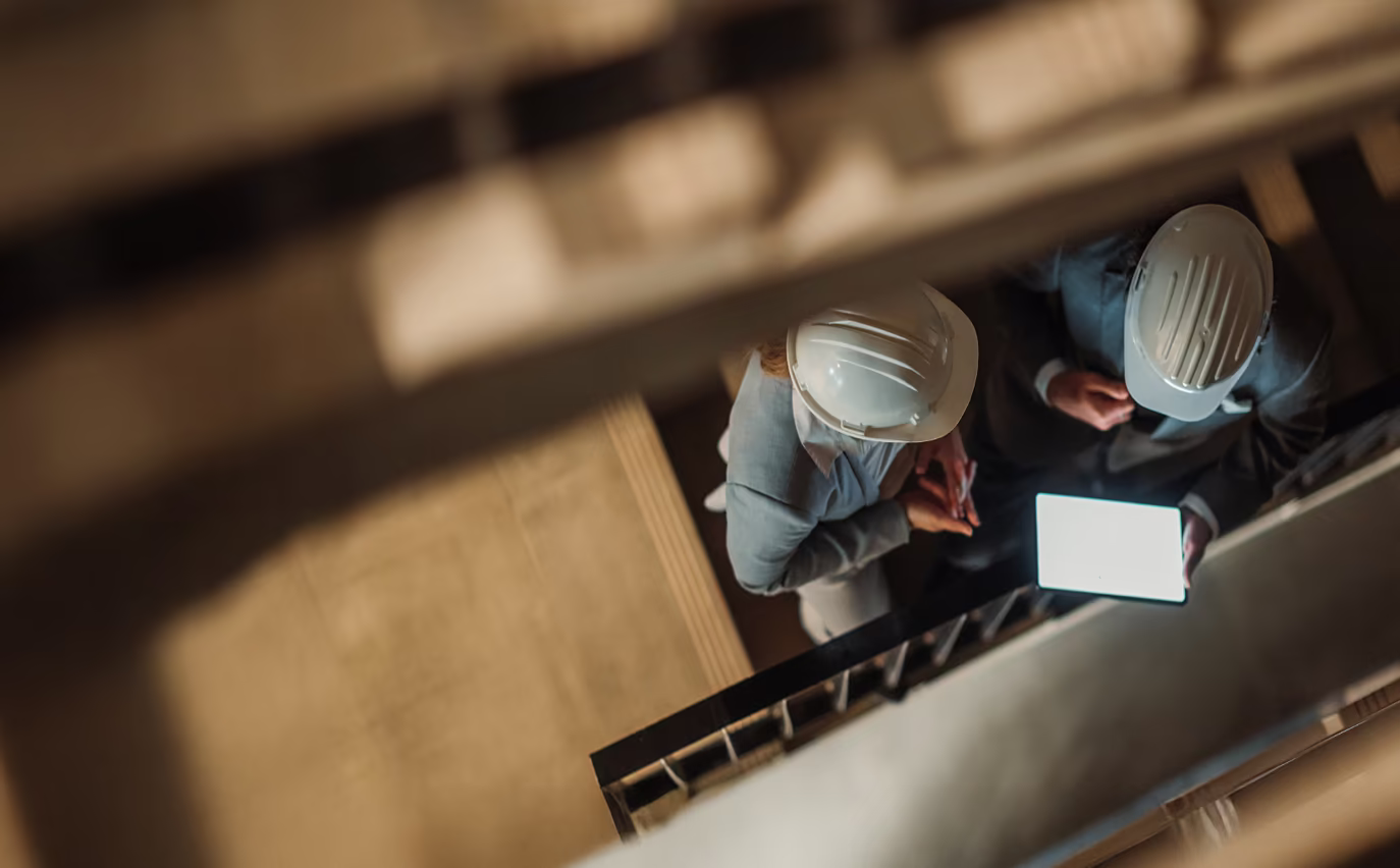 Dos profesionales de la construcción con cascos blancos revisando la pantalla de una tableta en una escalera.