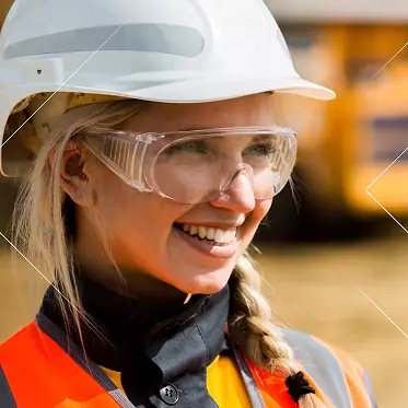 Obrera sonriente con casco blanco, gafas de seguridad transparentes y chaleco de seguridad naranja.