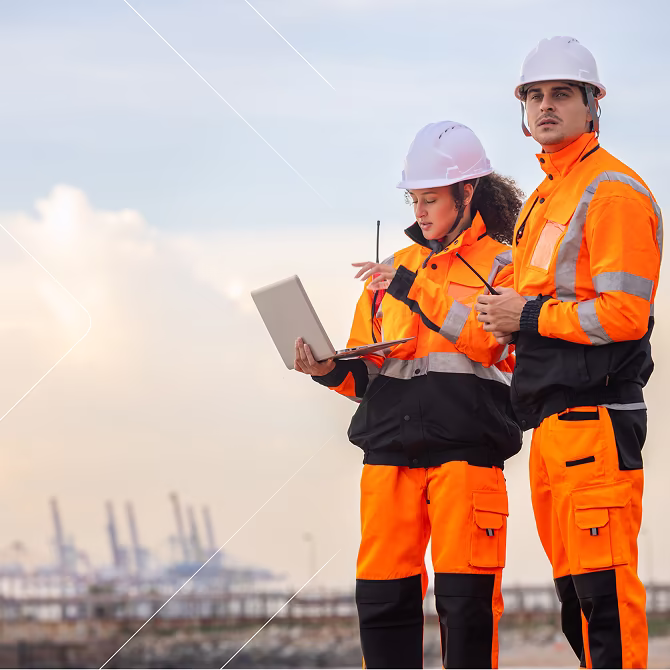 Dos trabajadores de la construcción con chaquetas naranjas de alta visibilidad y cascos blancos, uno con un ordenador portátil y el otro con un walkie-talkie, de pie al aire libre cerca de un sitio de trabajo industrial.