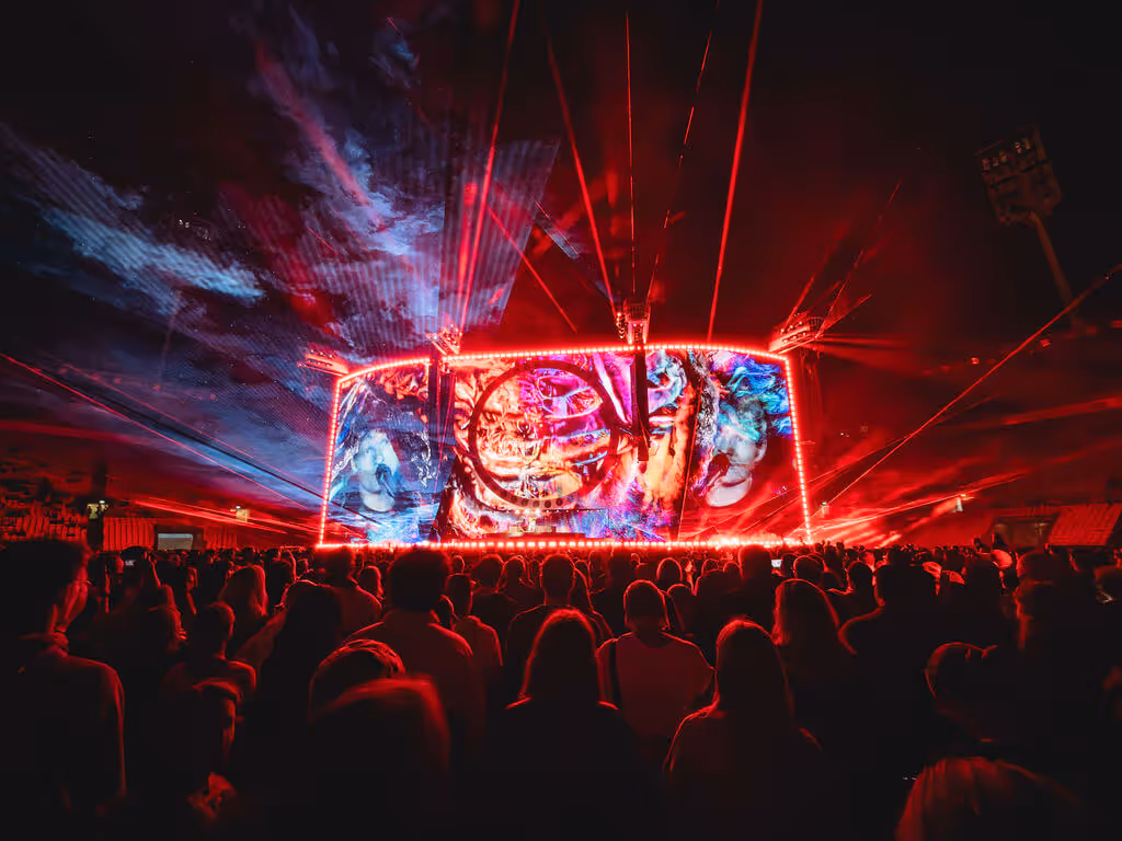 Crowd watching a large illuminated stage with colorful, abstract projections and red laser lights at night.