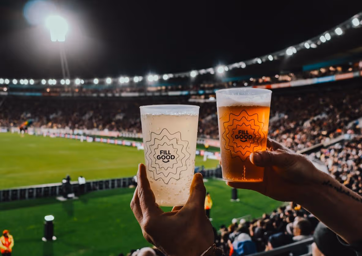 Two hands holding plastic cups with beverages labeled 'FILL GOOD' in a brightly lit stadium during an event.