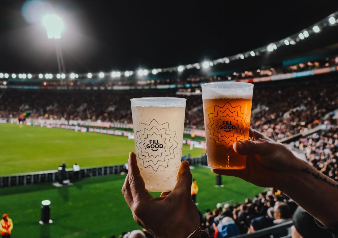 Two hands holding plastic cups with beverages labeled 'FILL GOOD' in a brightly lit stadium during an event.