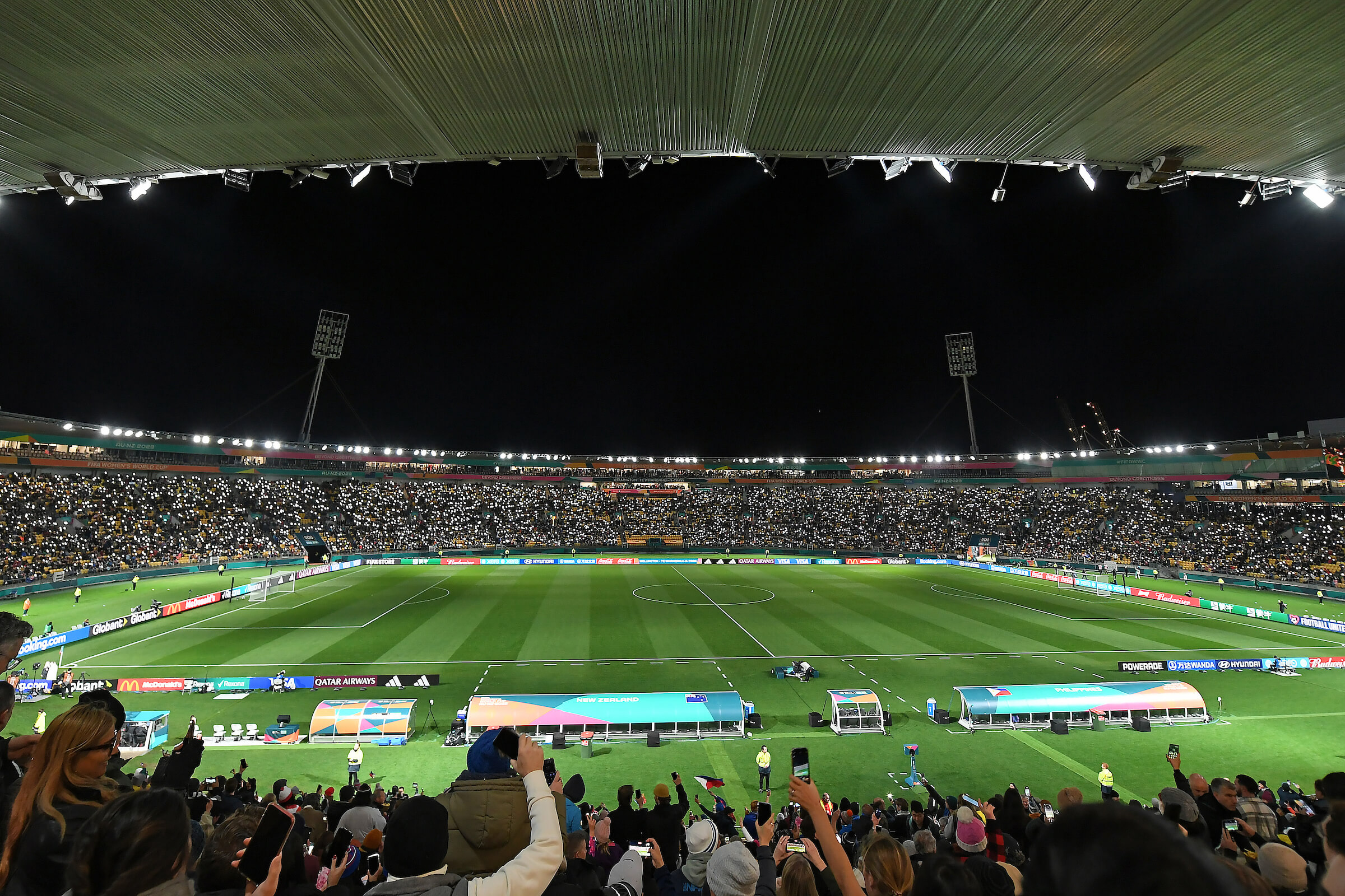 Night view of a football stadium filled with spectators holding up lit mobile phones, illuminating the stands around the field.