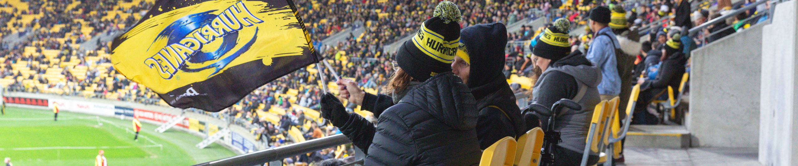 Fans in winter hats waving a yellow and black Hurricanes flag inside a stadium with a soccer field in the background.