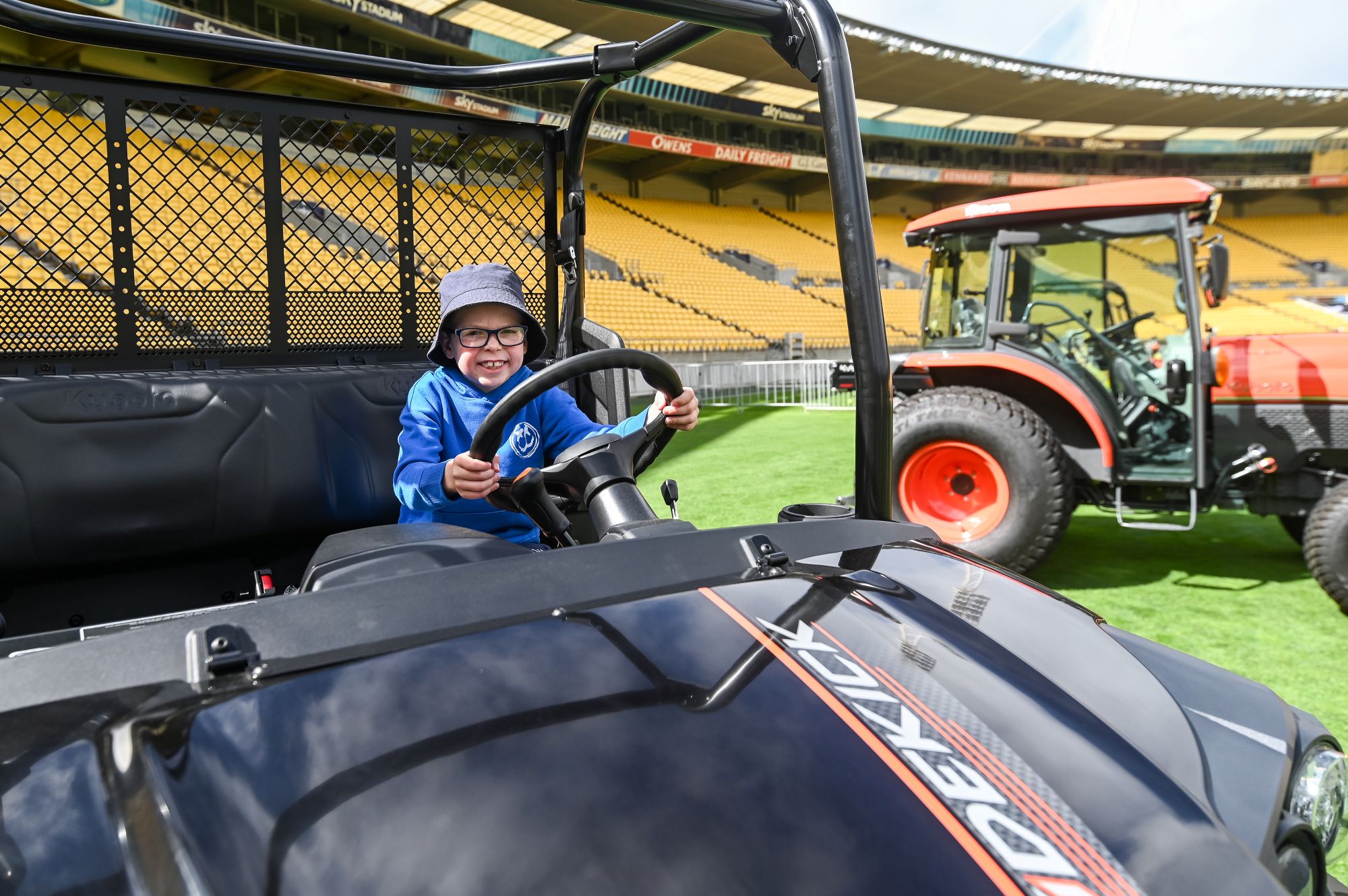 Smiling child wearing glasses and a bucket hat sits at the steering wheel of a Kubota utility vehicle on a sports stadium field.