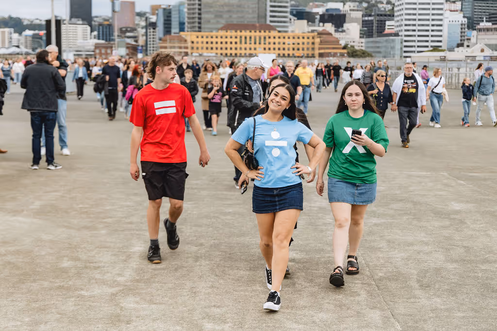 A young woman in a blue t-shirt and denim skirt walking and smiling, flanked by a man in a red t-shirt and shorts and a woman in a green t-shirt and denim skirt, with a crowd and city buildings in the background.