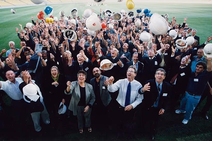 Group of diverse professionals standing on a field, joyfully throwing hard hats into the air in celebration.