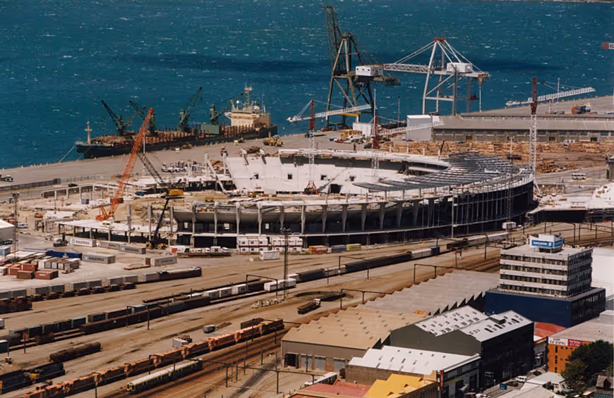 A large stadium under construction near a waterfront port with cranes, shipping containers, and train tracks in the foreground.