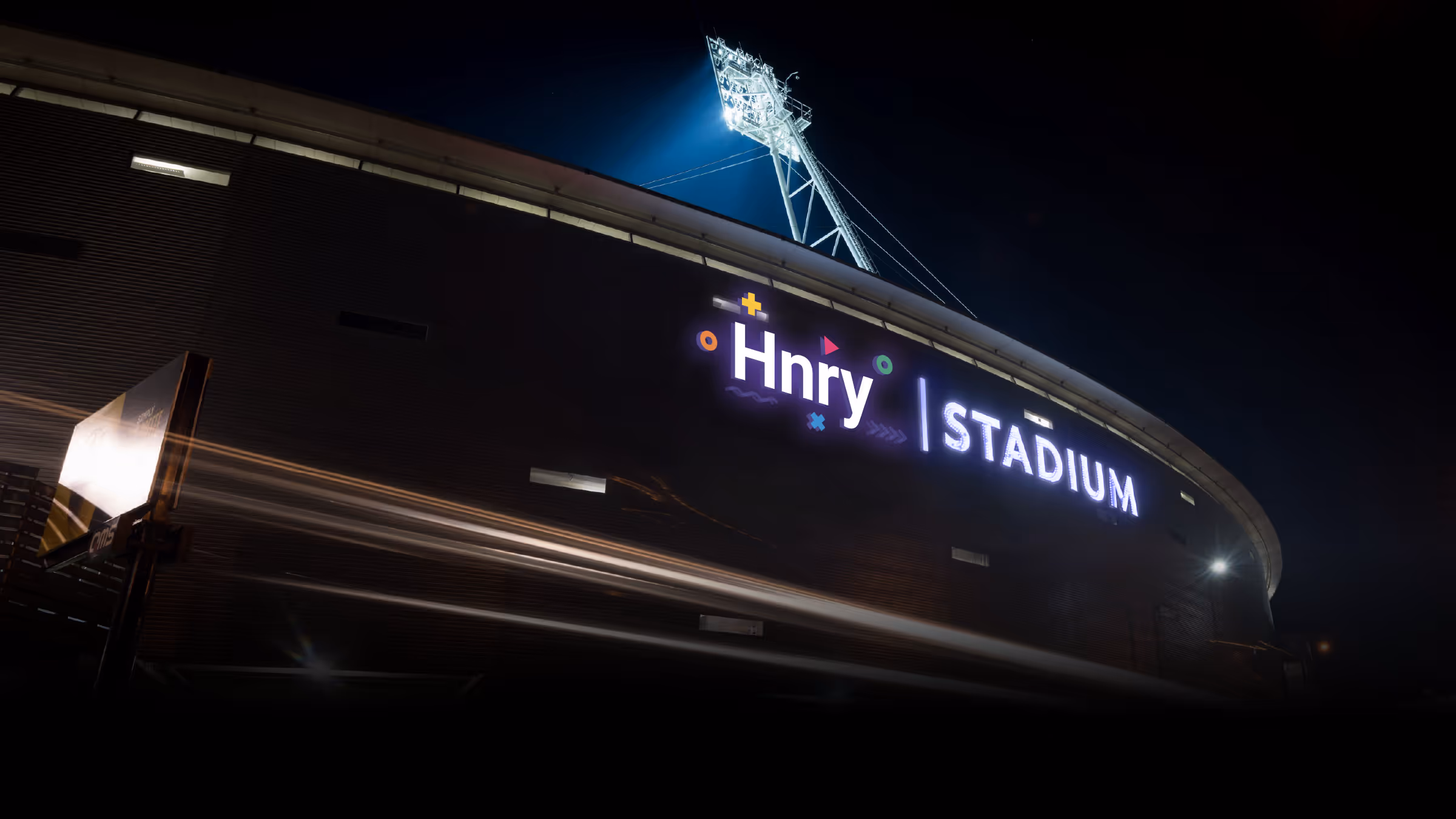 Night view of Hnry Stadium illuminated with bright signage and a tall floodlight tower.
