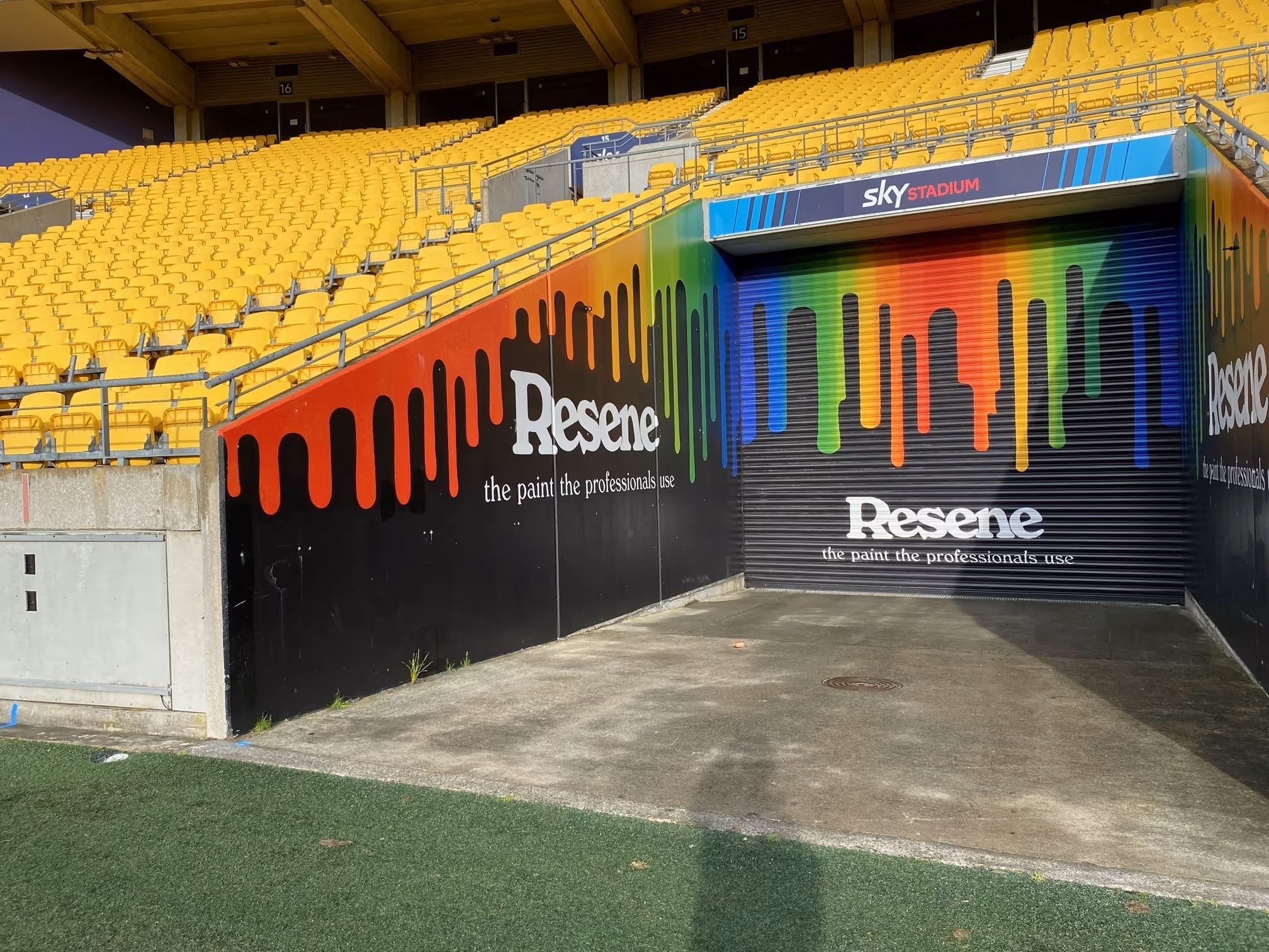 Empty yellow stadium seats above a black wall with colorful paint drips and the Resene logo with the tagline 'the paint the professionals use'.