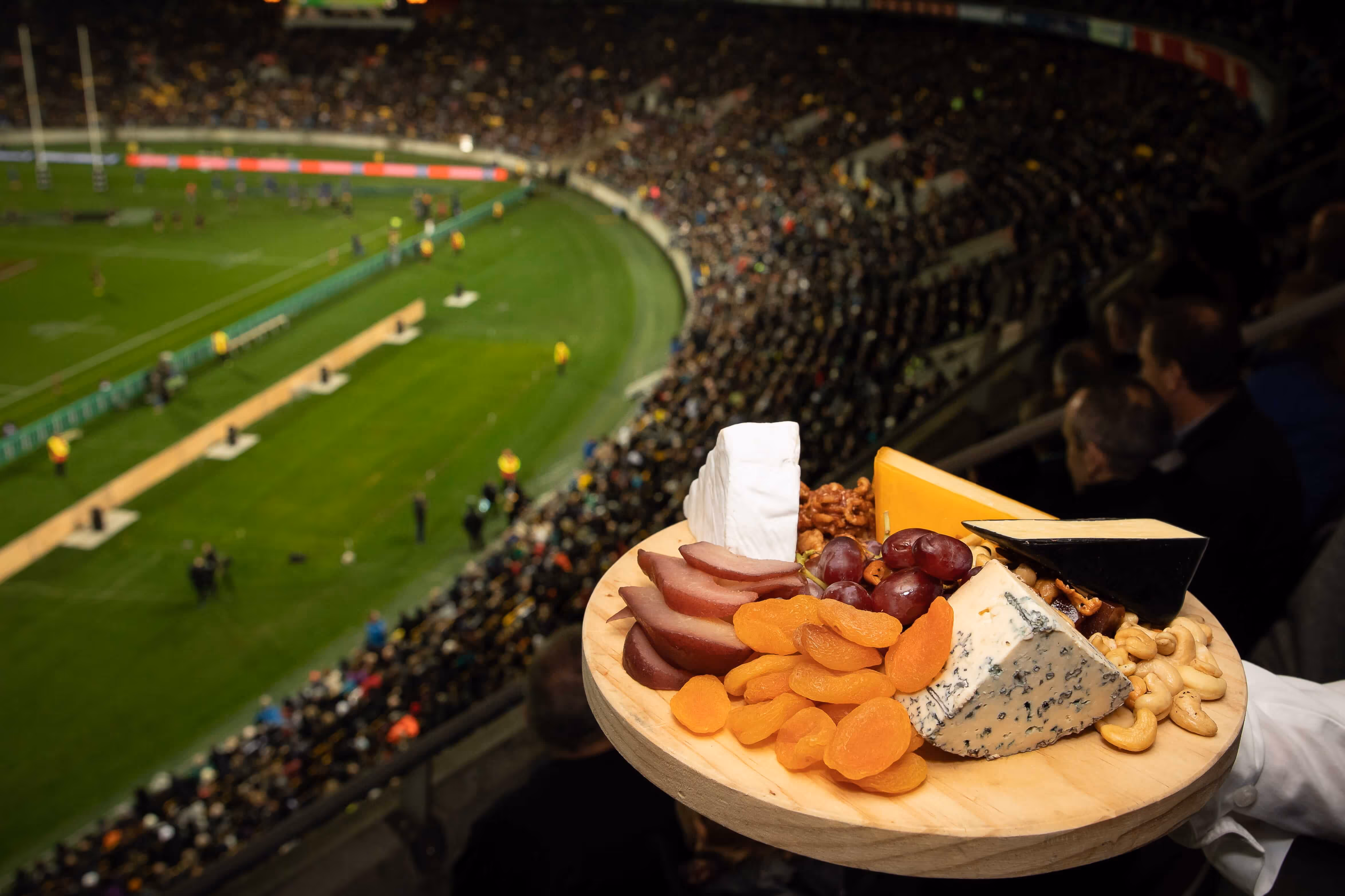 Wooden platter with assorted cheeses, dried apricots, grapes, nuts, and slices of fruit held above a crowded rugby stadium.