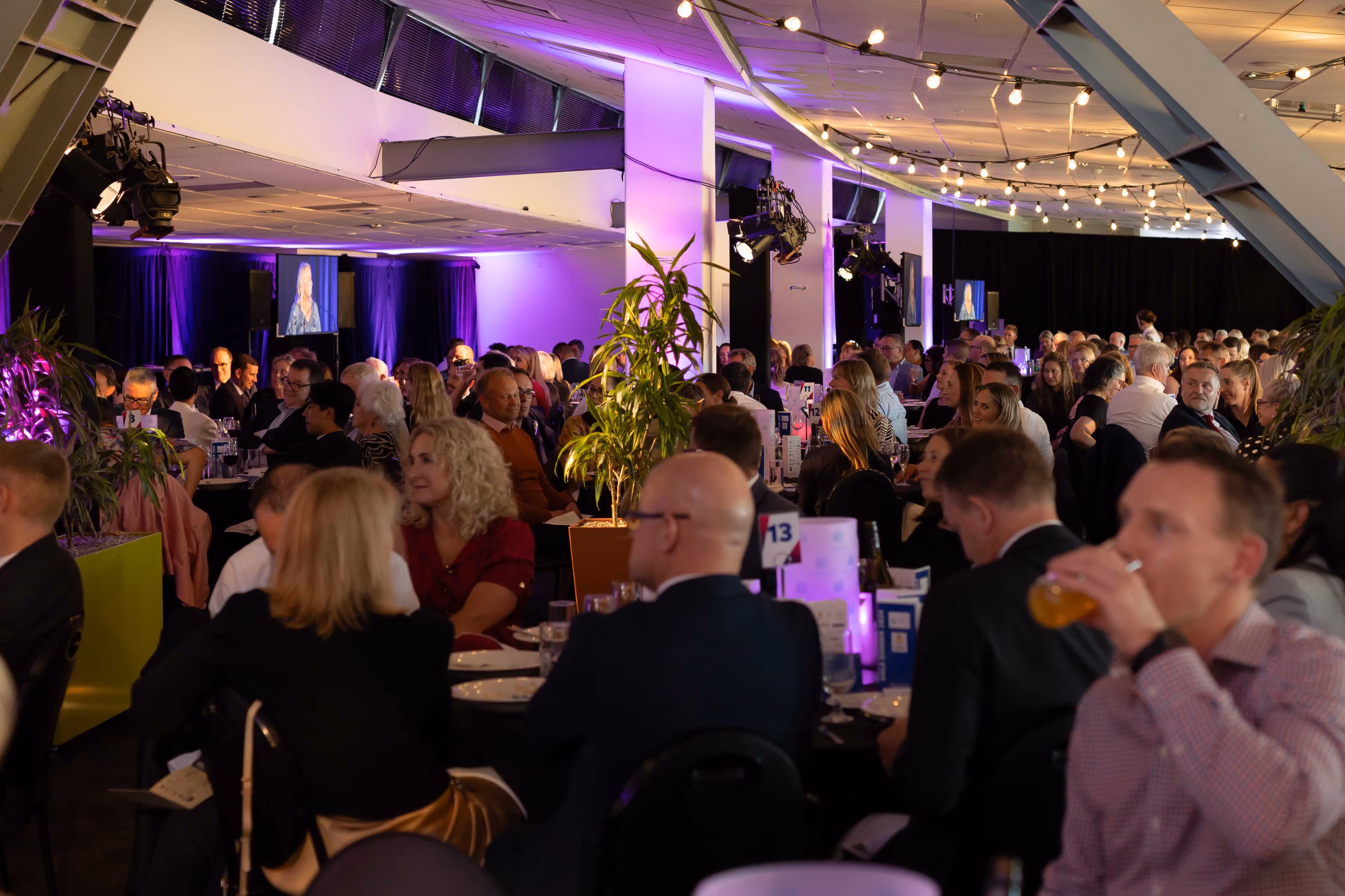 Large group of people seated at round tables in a dimly lit event space with purple lighting and string lights, watching a speaker on screens.