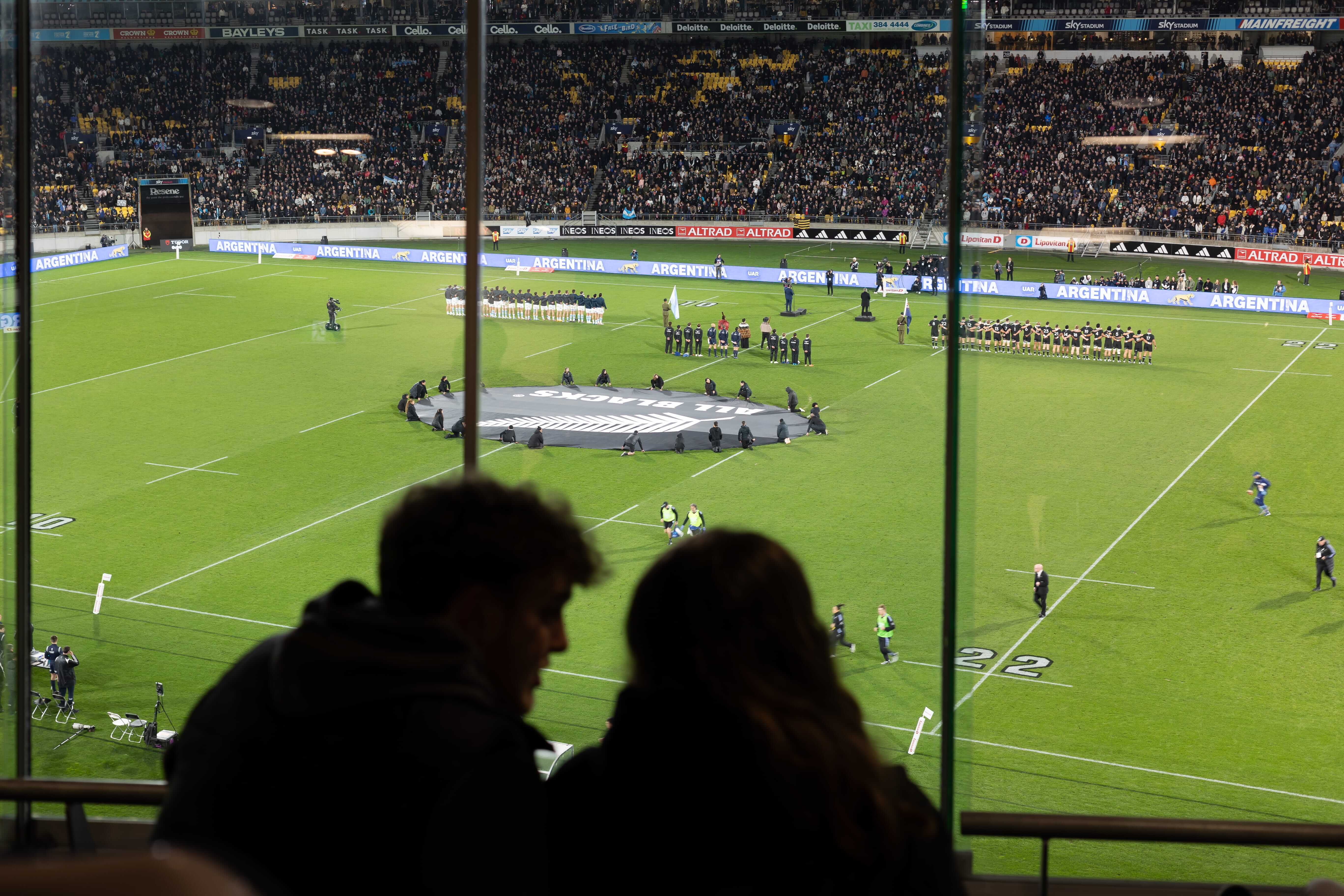 Rugby teams lined up on the field at a stadium with an All Blacks logo banner in the center and a large crowd in the stands.