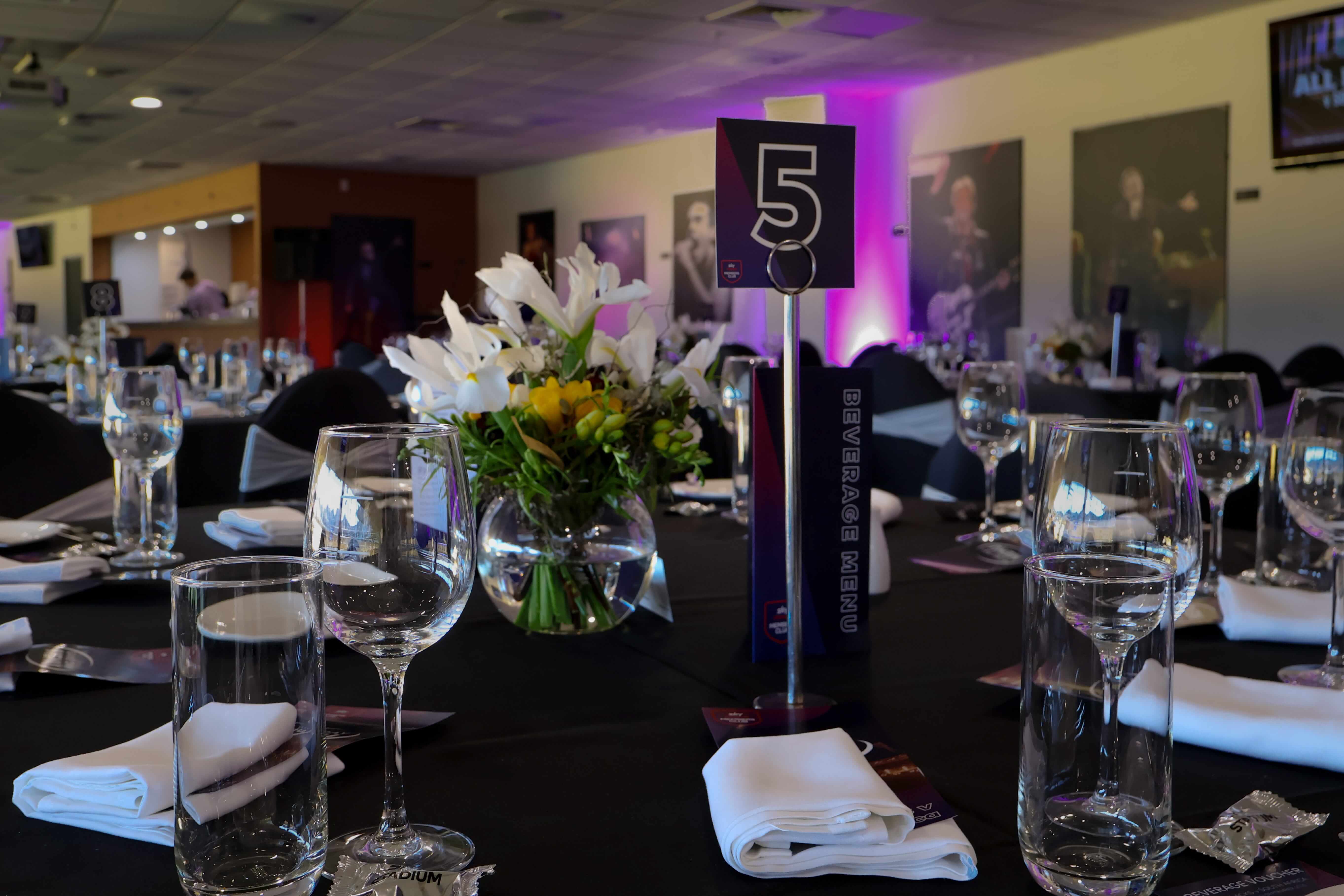 Table set for an event with glasses, white napkins, a floral centerpiece, and a table number 5 with a beverage menu.