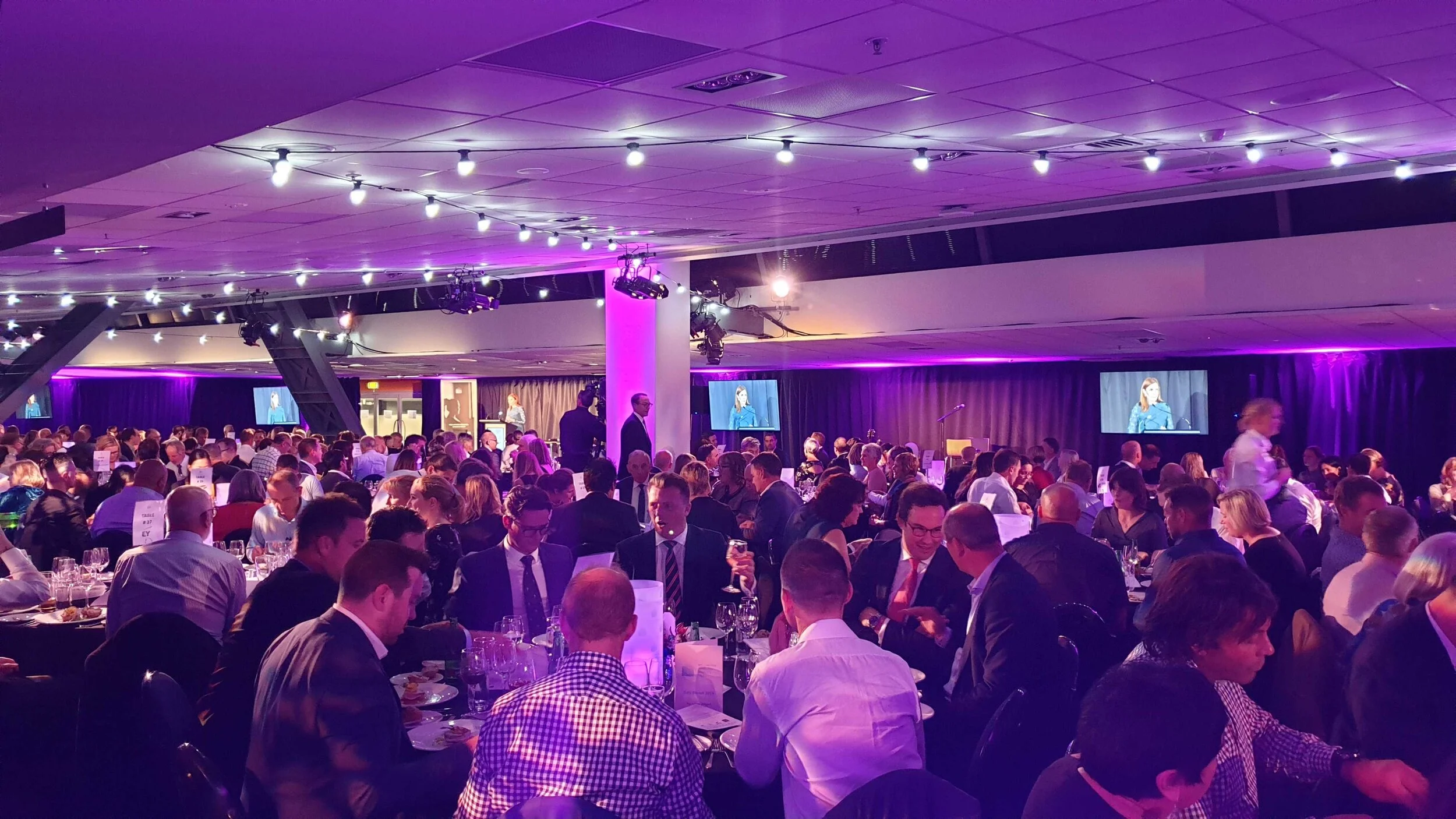 Large banquet hall filled with people seated at round tables dressed for a formal event with purple lighting and a speaker shown on screens.